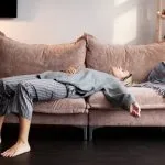 Woman in her 40s lying exhausted on a couch at home, illustrating early signs of perimenopause such as fatigue, sleep issues, and menopause transition symptoms. Solutions offered at Greenwich Medical Spa.