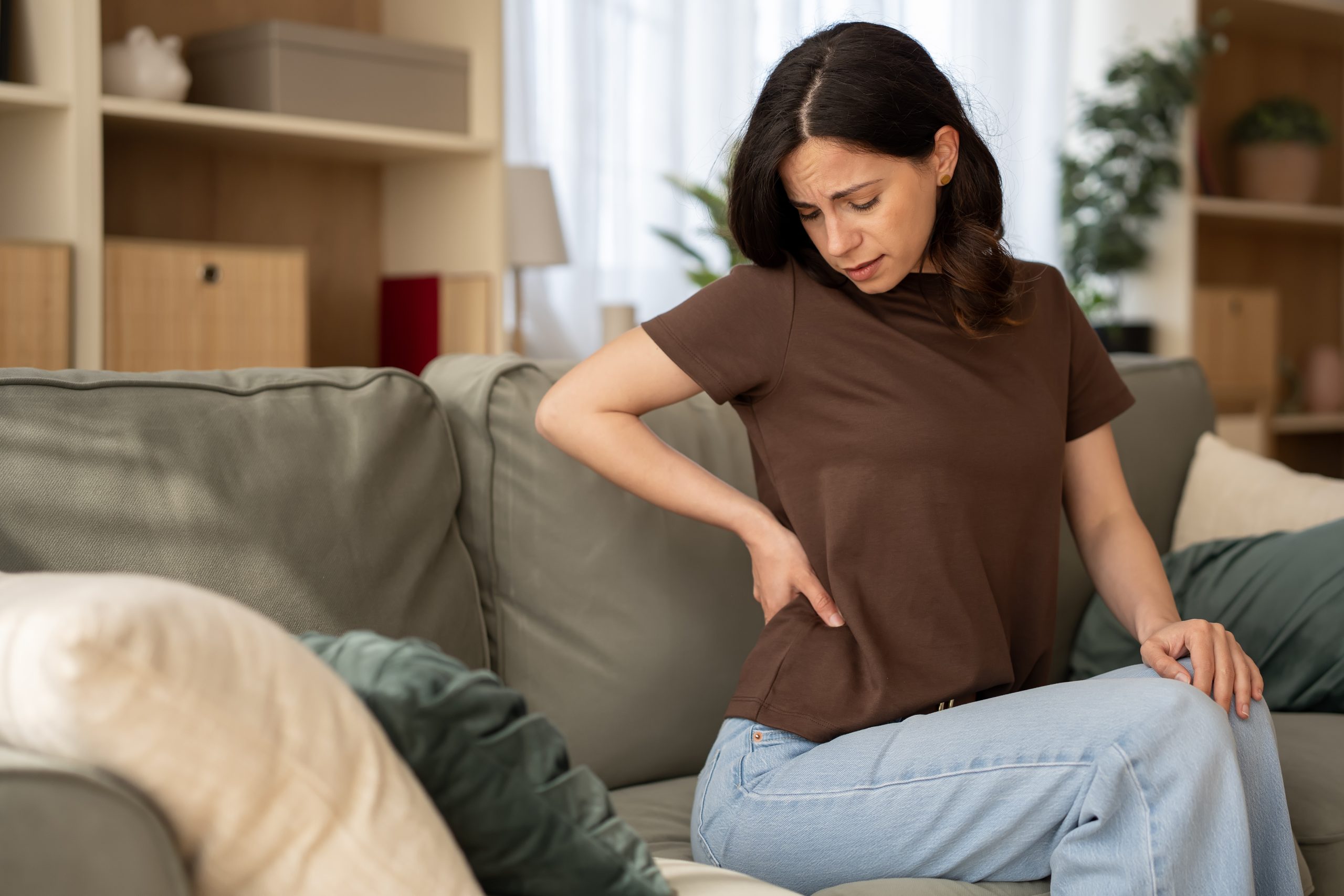 Woman,Sitting,On,A,Sofa,And,Holding,Her,Lower,Back A woman experiencing perimenopause joint pain and body aches, illustrating menopause symptoms discussed by Greenwich Medical Spa.